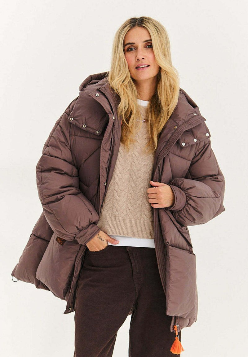 Brązowa puchowa kurtka z kapturem, z zatrzaskami i pomarańczowym suwakiem. Noszona na beżowym swetrze w warkocze i ciemnych spodniach.