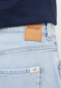 Jeans de mezclilla azul claro con una etiqueta de cuero marrón marcada "MUSTANG" y una pequeña etiqueta blanca con un logo en el bolsillo trasero.