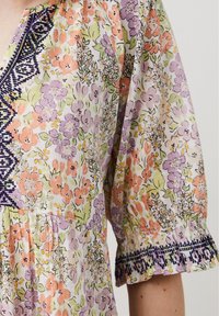 Blouse florale en tissu léger avec des fleurs pastel orange, violet et vert, accompagnées d'une broderie marine au niveau du décolleté et des poignets.