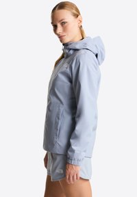 Γυναίκα με ξανθά μαλλιά σε αλογοουρά, ντυμένη με ανοιχτό μπλε ζακέτα με κουκούλα και ταιριαστό σορτς, που στέκεται πλάγια σε απλό φόντο.