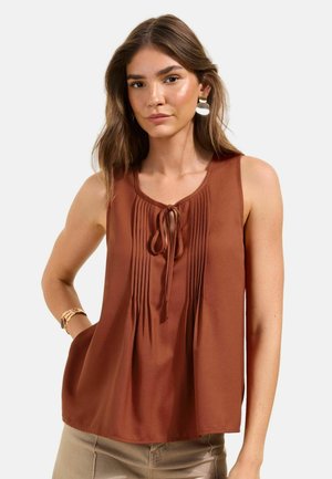 Hosszú barna hajú nő rust színű ujjatlan blúzt visel, elöl redőkkel és megkötővel, bézs nadrágot, arany órát és nagy fülbevalókat.