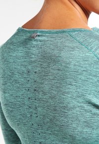 Lekki, długi rękaw koszulka w melanżowym odcieniu teal z okrągłym dekoltem i pionowym wzorem w kropki na plecach.