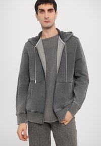 Grauer Zip-Hoodie mit weichem Material, zwei Vorderaschen, Kapuze mit Kordelzug und kontrastierendem grauem Innenfutter. Getragen über einem passenden Shirt.