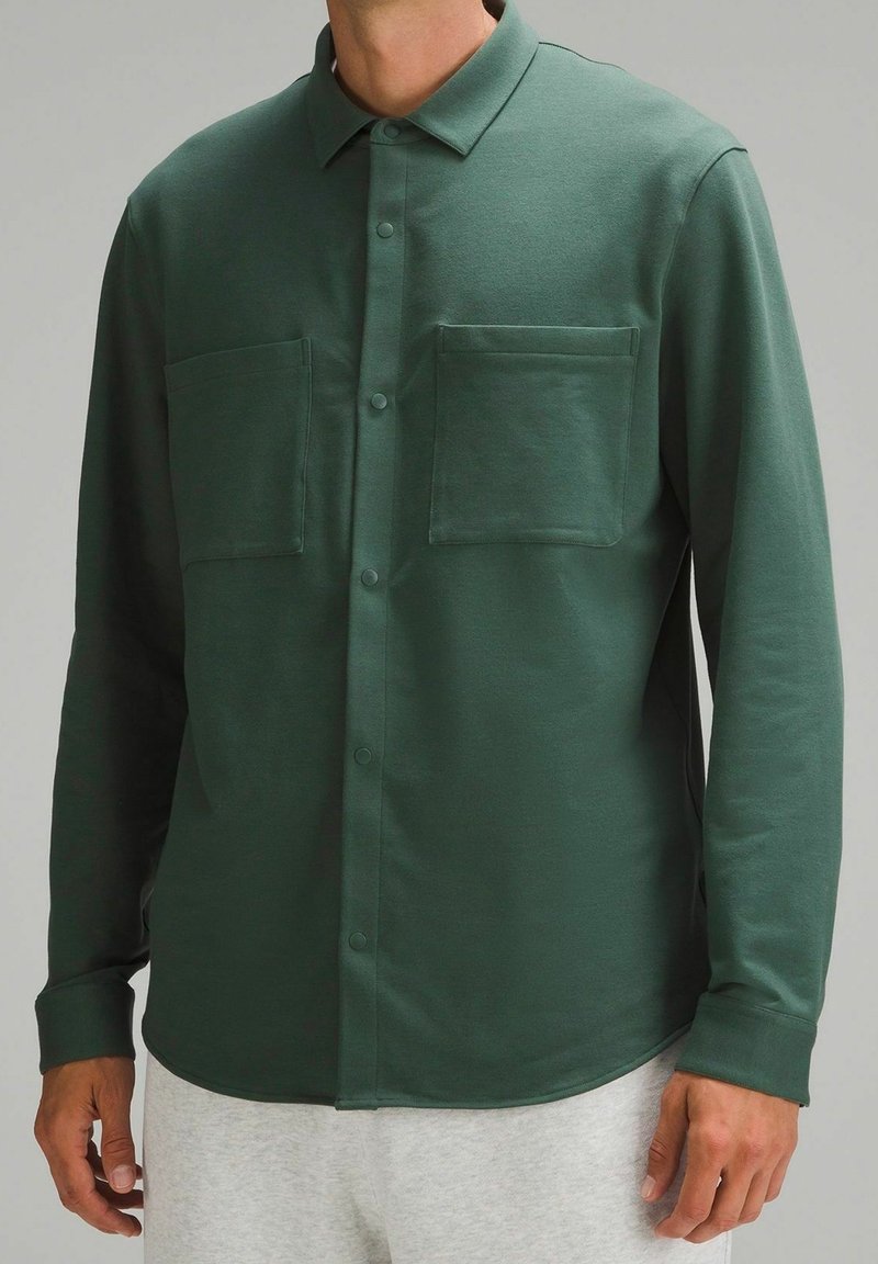 Camisa verde de manga larga en tela suave, con cuello tipo polo, botonadura frontal y dos bolsillos en el pecho con dobladillo redondeado.