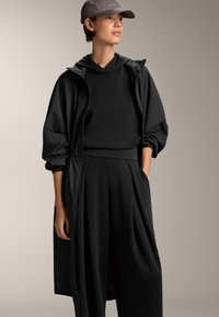 Felpa nera con cappuccio dal texture morbido, abbinata a un lungo cappotto nero con vita stretta e pantaloni neri larghi. Cappello grigio in accent.