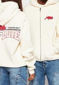 Roomkleurige hoodie met ritssluiting en voorkantzakken. Heeft rode geborduurde accenten en afbeeldingen op de achterkant, inclusief de tekst "LOVE PROJECT".