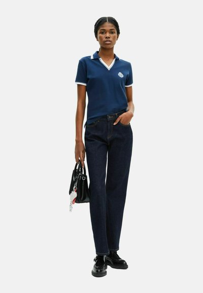 Marineblå polo t-shirt med hvid kant, parret med mørkeblå lige ben jeans. Modellen holder en sort håndtaske med en charm. Enkeltfarver, afslappet design.