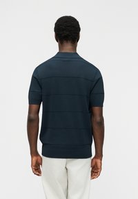 Άνδρας που φοράει σκούρο μπλε πόλο με κοντά μανίκια και διακριτικές οριζόντιες ρίγες, καθώς και ανοιχτόχρωμο παντελόνι, γυρισμένος με την πλάτη προς το φακό σε απλό φόντο.