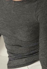 Närbild på en person som bär en mörkgrå ribbad stickad topp med lång ärm och lätt glans, ihop med ljusa byxor.