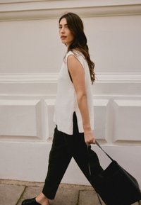 Pull en maille blanc sans manches avec fentes sur les côtés, associé à un pantalon noir et un sac cabas noir. Texture lisse avec des accents côtelés.