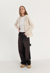Felpa zip up beige chiaro con cordini bianchi, abbinata a jeans neri larghi e zoccoli beige. Tessuto morbido, vestibilità rilassata, design semplice.