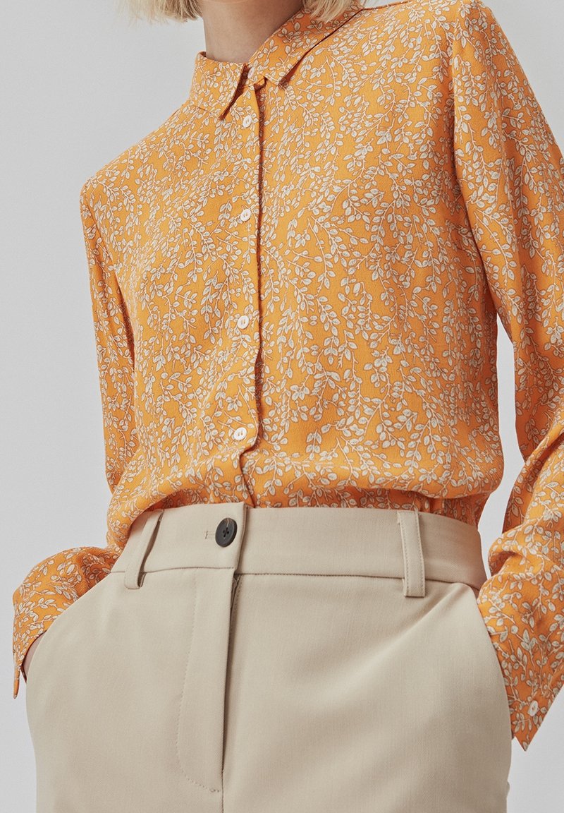 Pomarańczowa koszula zapinana na guziki z białymi kwiatowymi wzorami, długimi rękawami i spiczastym kołnierzykiem. Połączona z beżowymi spodniami z czarnym guzikiem.