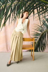 Mujer con falda plisada beige, chaleco blanco sin mangas con botones, tacones con estampado de cebra, apoyada en una silla de madera frente a un fondo de hojas de palma.
