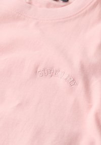 Jasnoróżowa bawełniana koszulka z okrągłym dekoltem, z haftowanym napisem "SUPERDRY" w zakrzywionym kształcie na klatce piersiowej.