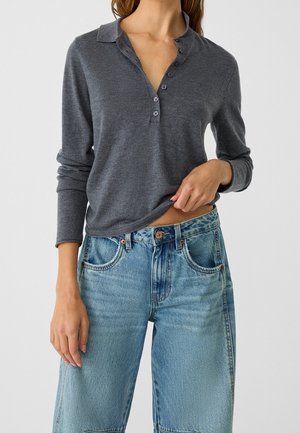 Vrouw draagt een donkergrijs langmouws henley-shirt en lichtblauwe high-waisted spijkerbroek, poseert met één hand waarmee ze de zoom van het shirt optilt.