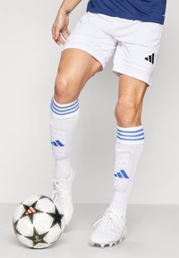 Shorts de sport blancs avec trois rayures noires, chaussettes rayées bleues et chaussures de football blanches. Un ballon de football noir et blanc avec des accents en forme d'étoile est à proximité.