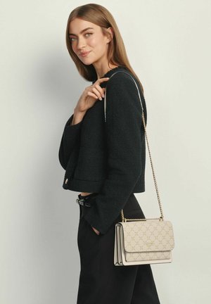 Kobieta o długich włosach, ubrana w czarną kurtkę i spodnie, trzyma beżową torebkę w wzory z złotym łańcuszkiem na ramieniu.