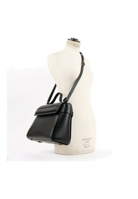 Sac à main en cuir noir avec bandoulière réglable et poignée supérieure présenté sur un mannequin de robe blanc.