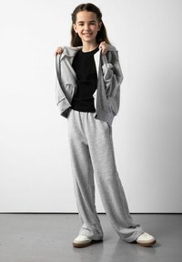 Jasnoszara bluza z kapturem, rozpięta, na czarnej dopasowanej koszulce; szare dresy, luźny krój; białe trampki z brązowymi podeszwami; jednolite tło.