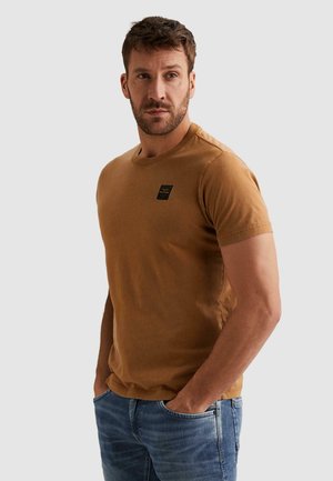 Man met baard, gekleed in een bruin T-shirt met korte mouwen en een blauwe spijkerbroek, staand met handen in de zakken, kijkt lichtjes opzij.