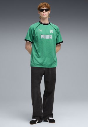 Jonge man die een groen Puma sportshirt met nummer 10 draagt, zwarte spijkerbroek, zwart-witte schoenen en donkere zonnebril, staand met handen achter de rug.