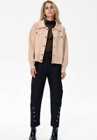 Beżowa, dziana kurtka z kołnierzykiem, czarnymi guzikami i dwiema kieszeniami na piersi; w komplecie z czarnymi spodniami z detalami guziczków na kostkach.