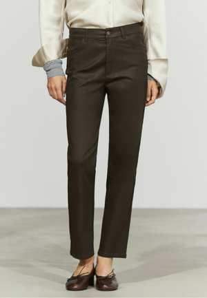 Vrouw draagt donkerbruine slim-fit broek met een crèmewitte blouse en bruine lage hakken en staat op een grijze vloer.