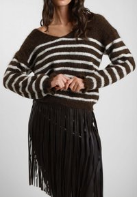 Maglione a righe marroni e bianche in maglia, con una scollatura ampia e dettagli in frange all'orlo, indossato sopra una gonna nera lucida con frange.