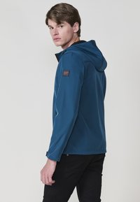 Giacca blu con cappuccio da uomo realizzata in tessuto morbido, con zip frontale, tasche laterali e un'applicazione con logo sulla spalla.
