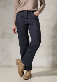 Jeans de mezclilla azul oscuro con un corte recto, con detalles de costura, cinco bolsillos y una textura sutil. Combinados con zapatos beige.