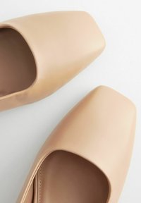 Neliöpäiset, pehmeästä beige nahasta valmistetut slip-on-kengät. Sileä pinta, jossa on vain vähän ompeleita ja helposti jalkaan laitettava muoto. Ei näkyviä metalliosia tai koristeita.