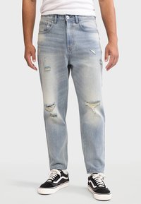 Jeans azzurri chiari con varie strappi, vestibilità dritta, texture sbiadita e chiusura con bottone in vita, abbinati a sneakers nere.