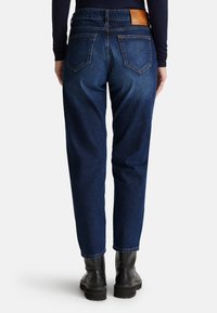 Jean en denim bleu foncé avec une coupe décontractée, couture visible, poches arrière angulaires et une étiquette en cuir beige sur la taille.