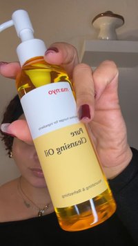 Bottiglia trasparente con un olio detergente giallo; erogatore a pompa bianco; etichetta con testo rosso su sfondo bianco e giallo.