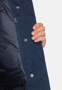 Marinblå quiltad jacka med dragkedja och tryckknappar, har en slät innerfoder och en hand som håller jackans kant.