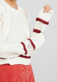 Weißer gestrickter Pullover mit offener Maschenstruktur und kastanienbraunen Streifen auf den Ärmeln. Verfügt über gerippte Bündchen und einen strukturierten Körper.