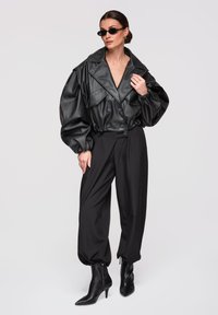 Vrouw draagt een oversized zwarte leren jas, zwarte broek met hoge taille, puntige laarzen met hak en donkere zonnebril tegen een effen achtergrond.