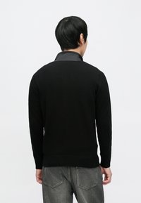 Juodas megztinis su apvalia apykakle ir raišteliais rankogaliuose, dėvimas ant pilko pūsto striukės, pasižymintis lygiu mezgimo paviršiumi ir aptemptu dizainu.