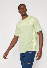 Αθλητική κοντομάνικη μπλούζα ανοιχτού πράσινου με υφή υφάσματος, λείο μέσο μέρος και διακριτικό γκρι λογότυπο, φορεμένη με μπλε cargo σορτς.