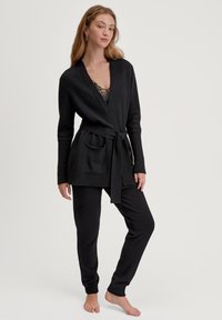 Vrouw die blootsvoets staat, gekleed in een zwarte gebreide wikkelcardigan met een riem en bijpassende gebreide broek, met een kanttop die eronder zichtbaar is, op een neutrale achtergrond.