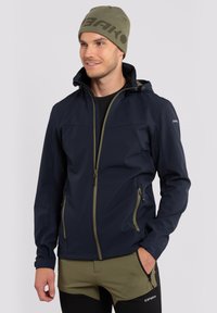 Marineblauwe softshell jas met groene accenten, voorrits en capuchon. Gecombineerd met olijfgroene shorts, voorzien van zijzakken en logo-opdruk.