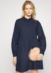 Vestido azul-marinho de manga longa com fecho de botão, colarinho e faixa elástica na cintura. Modelo segura uma clutch rosa fofinha. Tecido de algodão suave.