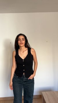 Γυναίκα με μακριά σκούρα μαλλιά που φοράει μαύρο αμάνικο τοπ με κουμπιά και σκούρο τζιν, στέκεται μέσα σε έναν χώρο μπροστά σε έναν απλό τοίχο.