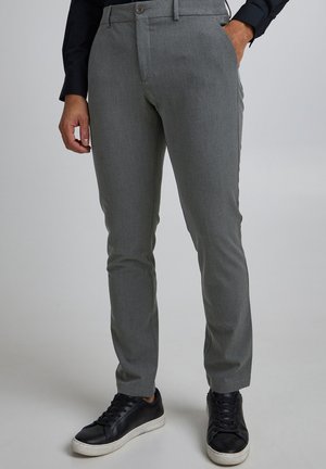 Mann trägt slim-fit graue Anzughose mit Gürtelschlaufen, kombiniert mit schwarzen Turnschuhen und einem schwarzen Langarmhemd, eine Hand in der Tasche.