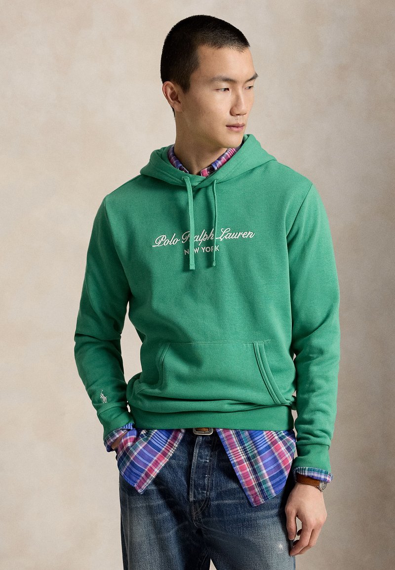 Νέος άνδρας φοράει πράσινο φούτερ Polo Ralph Lauren πάνω από μπλε και κόκκινο καρό πουκάμισο, με τα χέρια στις τσέπες, κοιτάζοντας προς τα πλάγια.