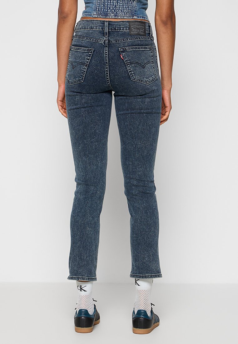 Femme portant un jean taille haute bleu foncé, un crop top bleu, des chaussettes blanches Calvin Klein et des baskets noires, se tenant contre un fond blanc uni.