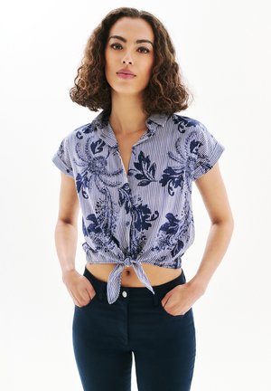 Chemise à manches courtes rayée bleu et blanc avec imprimé floral, nouée à la taille, dotée d'un col et d'une fermeture à boutons, associée à un jean noir.