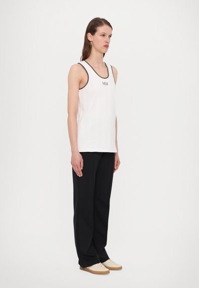 White ribbed tank top with dark trim, featuring "MM" logo, paired with loose black trousers and light-colored shoes.