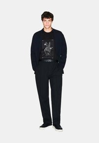 Schwarzes Graphic-T-Shirt mit weißem Friedenzeichen-Design, unter einem marineblauen Cardigan getragen. Schwarze Hose und schwarze Sneaker mit weißen Sohlen vervollständigen den Look.