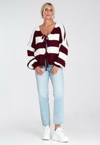 Kobieta w białej i burgundowej pasiastej kardiganie związaną z przodu, w jasnoniebieskich dżinsach, w białych platformowych sneakersach, stojąca na jednolitym tle.